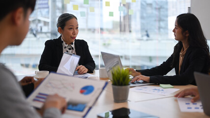 Experienced senior female team leader talking, giving instructions to diverse staff at corporate office