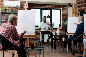 Man teacher explaining graphic illustration technique to students during art courses in creativity studio. Multiethnic team drawing vase sketch on white canvas learning new sketching skills