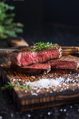 Medium-rare sliced beef steak with salt and herbs on a wooden board on a dark background.
