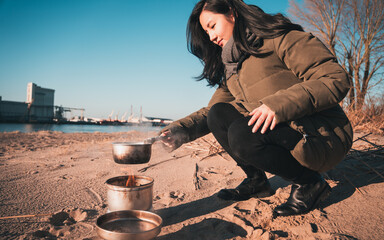 Young asian woman with warm clothes on is cooking with outdoor cooker on Harriersand beach at a riverbank during golden hour. Weser river blurred in the background.  - obrazy, fototapety, plakaty