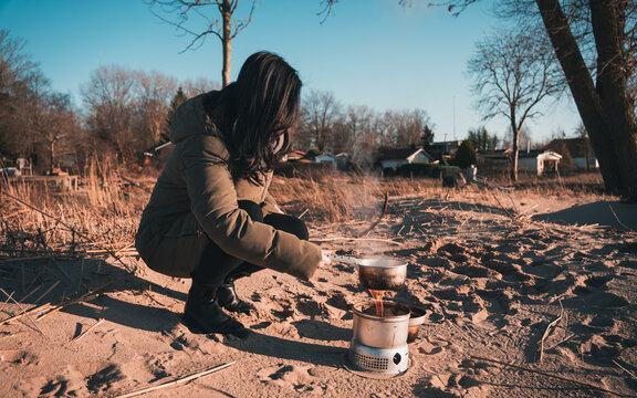 Young asian woman with warm clothes on is cooking with outdoor cooker on Harriersand beach at a riverbank during golden hour. Weser river blurred in the background. 