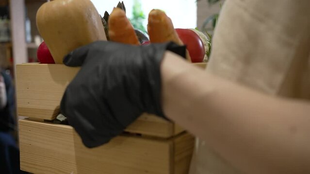 Woman in black rubber gloves carries wooden box filled with fresh vegetables. Cafe kitchen worker carries box with organic vegetables closeup