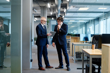 Obraz premium Mature businessman in suit showing something on digital tablet to his colleague while they standing at office