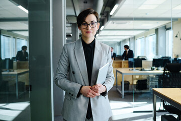 Portrait of successful young businesswoman in eyeglasses and formal wear looking at camera standing with tablet pc at office