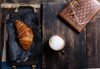 Croissant on a wooden table with a cup of coffee, antique knife and book.