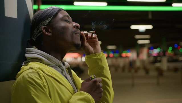 Black skin teenager takes a cigarette and lights it. Young African-American man smokes cigarette against background of evening city. Tired black man smokes marijuana against background evening lights.