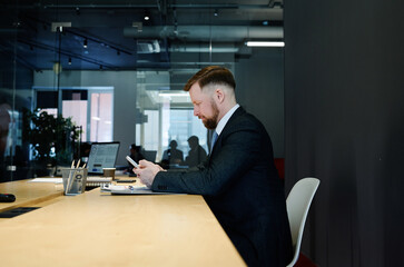 Young businessman in formal suit sitting at table in boardroom and using mobile phone awaiting meeting