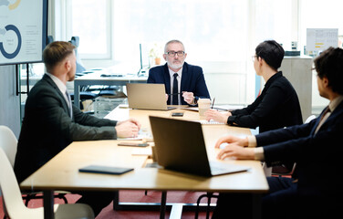 Senior manager in suit talking to partners at table with laptops during meeting at meeting room