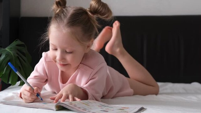 Portrait of a schoolgirl solves children crosswords on the bed. Logic games at home. Children solves crossword