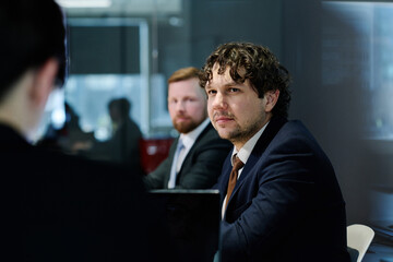 Young businessman in dark suit listening to speaker while sitting at table with his colleagues at meeting