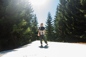 Stylish woman in casual clothes standing in the snow in the woods on a sunny spring day, walking on a mountain resort.