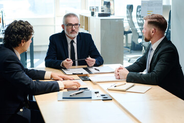 Group of serious businessmen in suits sitting together at table at boardroom and planning work in team