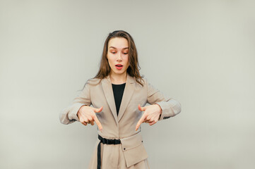 Positive businesswoman in a suit stands on a beige background and shows fingers down on an empty spot with a surprised face.