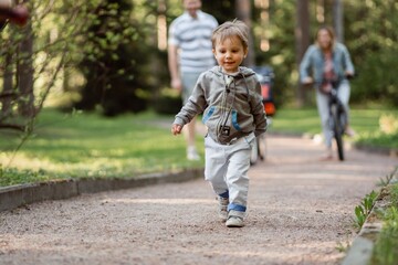 Fototapeta premium Family walking in park. Little boy on foreground, father and mother cycling on bicycle on background