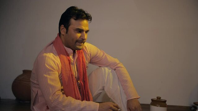 Frustrated village man smoking alone sitting at home - stressed lifestyle. Medium shot of a dissatisfied Indian villager wearing a Kurta-Pajama sitting in anger 