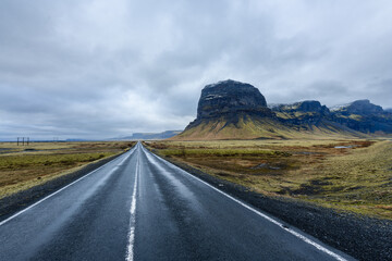 Straight road accross green barren scandinavian landscape with mountain ridge and peaks, cloudy sky