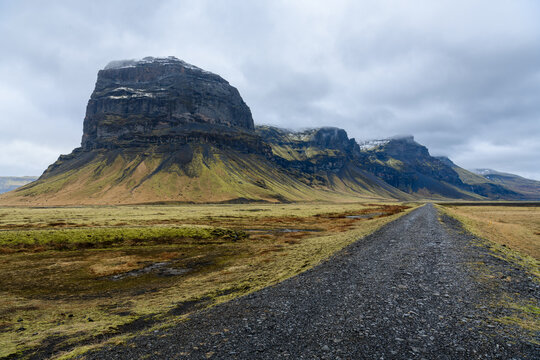 Straight gravel path accross green barren scandinavian landscape with mountain ridge and peaks, cloudy sky