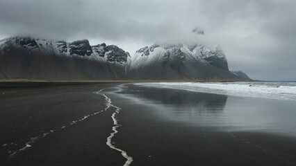 Black-sand beach coastline, white line of sea froth, background snow covered mountain, grey sky