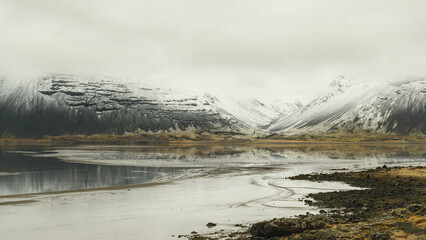Stony yellow-black lava lake shore, creek and lake, background snow covered black  mountains disappearing in white foggy sky