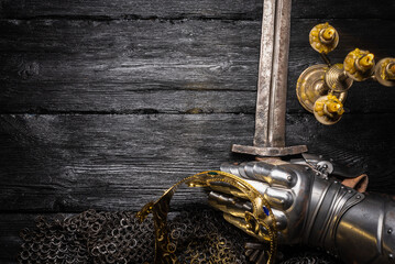 Ancient golden crown, armor gloves and knight sword in the light of burning candle on the wooden...