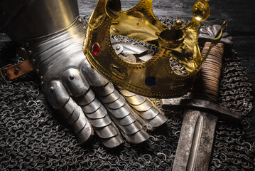 Ancient golden crown, armor gloves and knight sword on the wooden table close up background.