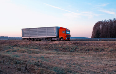 A semitrailer tractor with a tilt semitrailer transports cargo against the backdrop of an evening sunny sunset in summer. The concept of the logistics system and the business of cargo transportation