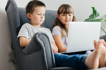 A schoolgirl with her brother are watching studying on a laptop while sitting in an armchair. Children are learning remotely online. Use of technology in the modern world.