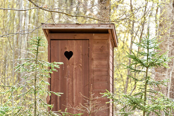Wooden outdoor toilet with heart on the door. Spruce trees around.