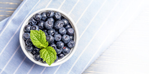Wild forest blueberries with fresh mint leaf in white bowl on blue striped napkin. Banner. Top view.