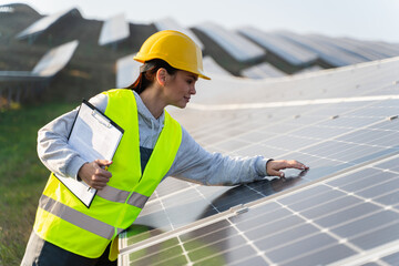 Caucasian female worker in signal vest touches solar panel and checks for damage. Concept of work at eco-friendly enterprise