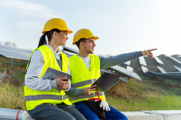 Portrait of engineers spending time outside near solar panels. Two workers: woman and man in special uniform pointing at something and smiling