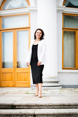 Beautiful happy woman in a black dress and white jacket walks through the park