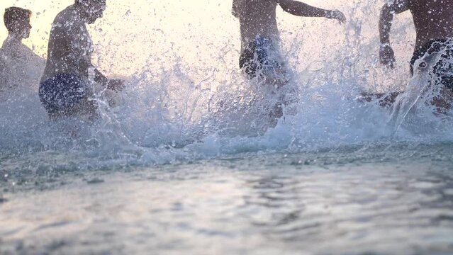 Group of happy friends at sunset running into water getting wet and making splashes swimming in the sea