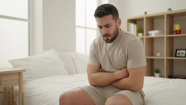 Young hispanic man suffering for stomachache sitting on bed at bedroom