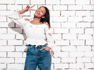 Portrait of young beautiful black woman. Smiling model dressed in summer jeans clothes. Sexy carefree female posing near white brick wall in studio. Tanned and cheerful. Shows peace sign and tongue