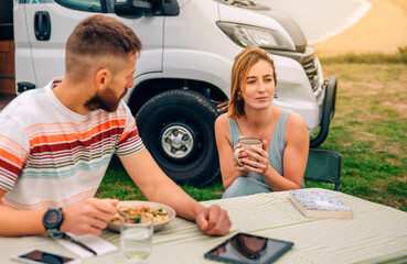 Young man looking to pensive woman holding coffee cup with camper van and beach on background