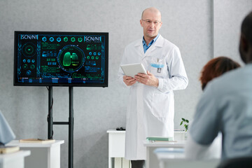 Mature lecturer in white coat with digital tablet standing near the display with human lungs and...