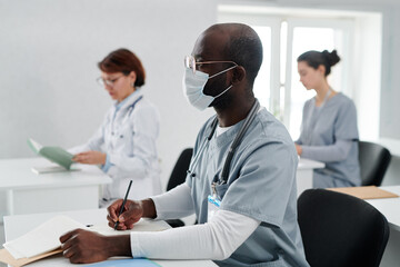 African young doctor in uniform and mask making notes in notebook at desk during medical training...