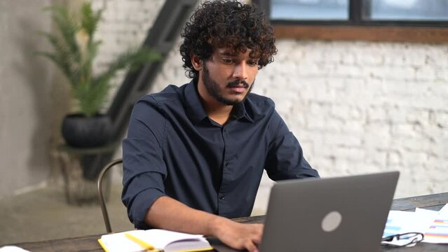 Modern bearded man working with laptop in home office. Indian male in smart casual shirt watching webinar and writing down, taking notes with a pen, planning schedule