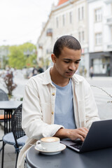 concentrated african american freelancer using laptop near cappuccino on cafe terrace.