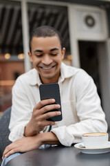 cheerful african american man texting on smartphone near cup of cappuccino on table on cafe terrace.