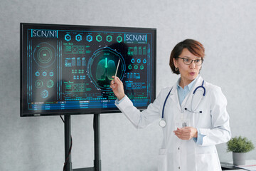 Mature female doctor in white coat pointing at digital display with chart and talking about CPR at...