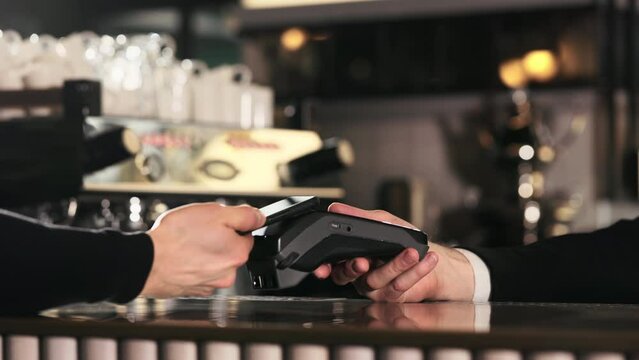 Close up of male client using modern smartphone for making cashless payment for drinks at car. Bartender standing behind counter and holding terminal.