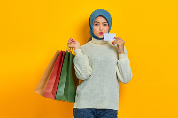 Cheerful beautiful Asian woman in white sweater holding shopping bags and credit card isolated over yellow background