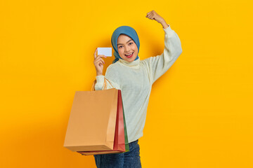 Cheerful beautiful Asian woman in white sweater holding shopping bag and credit card, celebrate luck isolated over yellow background