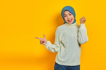 Excited beautiful Asian woman in white sweater pointing fingers aside at copy space isolated over yellow background