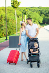 happy young parents mom and dad with a red suitcase and a baby in a stroller are going on a vacation trip in the summer, smiling