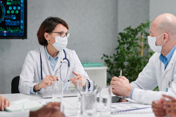 Two colleagues in masks and white coats discussing together at table during meeting at hospital