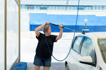 woman with short hair soaping a white car with a hose from a self-service station.