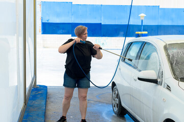 Adult woman with short hair cleaning a car with a pressure hose. Horizontal image.